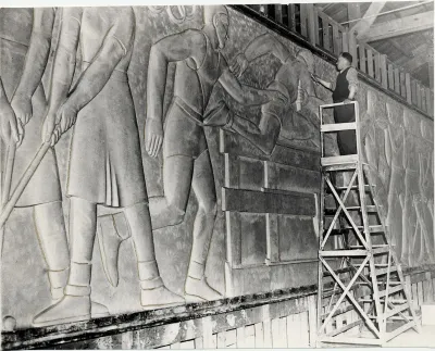 A Black man on a scaffold attending to a frieze-like artwork showing athletes running and jumping.
