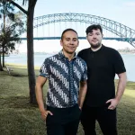 A brown man in a patterned shirt and a white man in a black shirt stand in a green park with a bridge behind them.