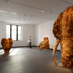 A person walking through a sunlit gallery with three abstract sculptures formed from carved wood.