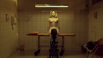 A half-nude person sitting on an operating table in a tiled room beneath a light. They stare at an animatronic creature with a human-like head.