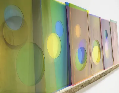 Multicolored glass panes layered atop one another and leaned against a white wall. Some have circular holes cut out of them that reveal unlike colors.