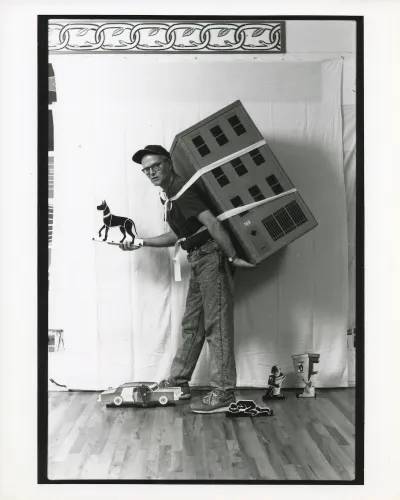 A man with a model of an apartment building strapped to his back holding a cut-out of a dog.