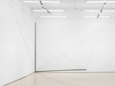 A sculpture composed of lines of barbed wire that hang from two walls and meet at a bar hanging above the floor.