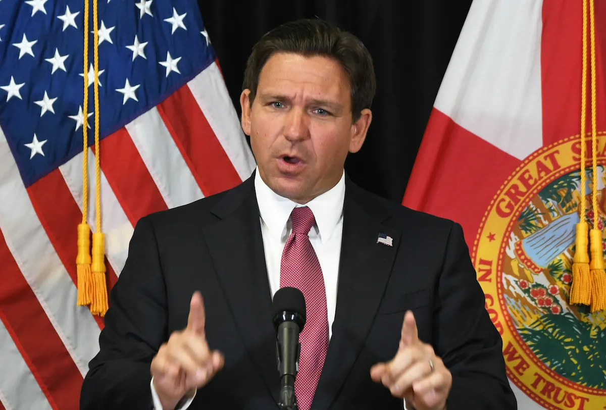 A white man holding up his two pointer fingers and standing before an American flag at a podium with a microphone.