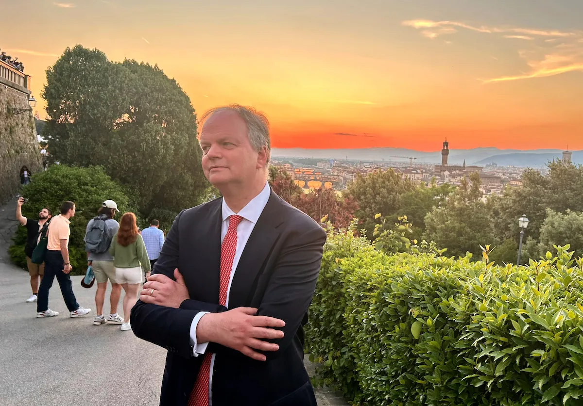 A white man in a suit and tie standing on a hilltop before a city at sunset. He crosses his arms and looks off into the distance.