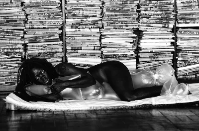A nude Black person crouched on the floor with plush covering their private parts. Stacks of newspapers can be seen behind them.