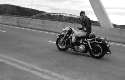 A black-and-white image of a man driving a motorcycle across a bridge and looking backward.