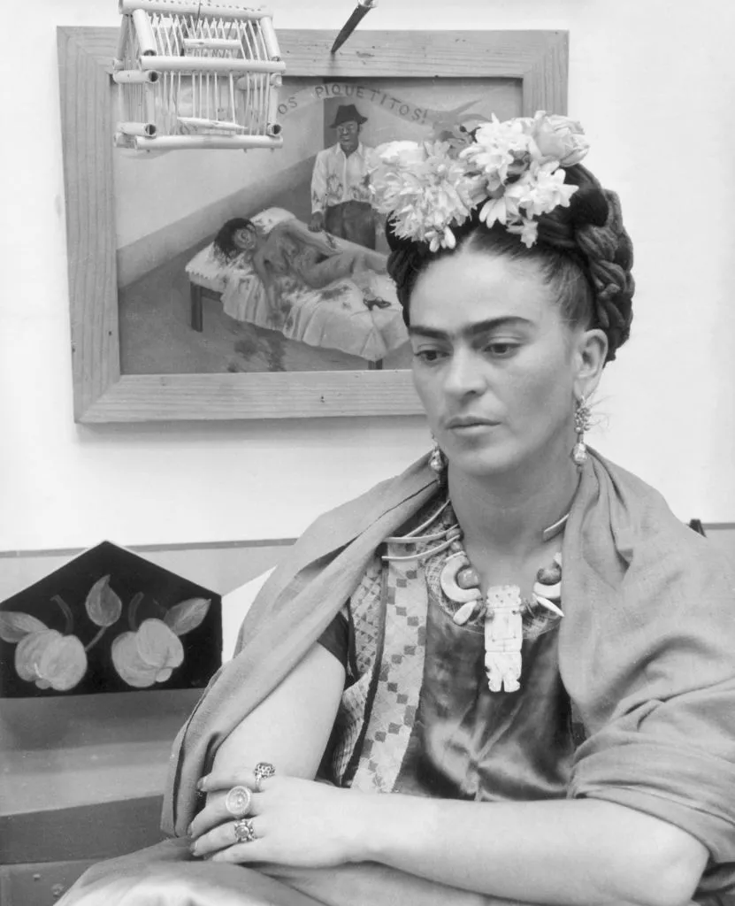 circa 1945:  Mexican painter Frida Kahlo (1907 - 1954) sits with her arms folded, looking down, in front of one of her paintings and a wooden bird cage. She wears flowers in her hair and a wooden necklace.  (Photo by Hulton Archive/Getty Images)