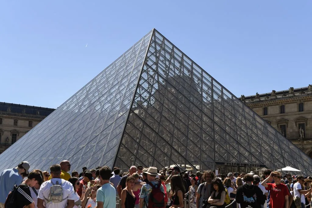 A crowd of people outside next to a large glass pyramid structure against a blue sky.