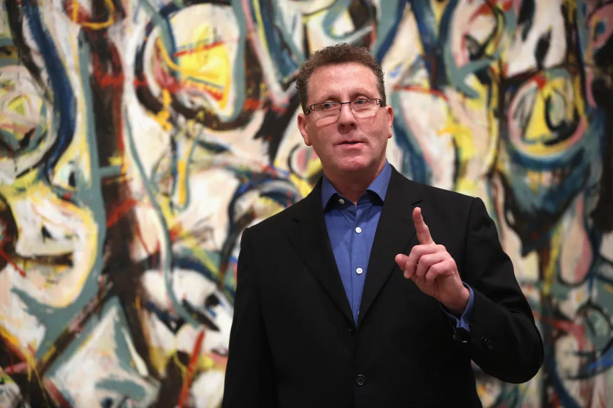 A white man in a blue shirt and black jacket raising one finger before a large abstract painting.