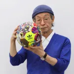 An Asian man in a blue cap and a blue cardigan holding a soccer ball with the Adidas logo and phantasmagorical imager.y