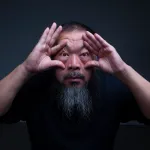 A portrait of an Asian man with a long beard spreading open the folds around his eyes.