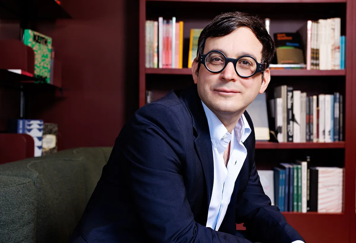 A white man in glasses wearing a blue suit and smiling on a chair with his hands on his lap. Behind him is a bookshelf.