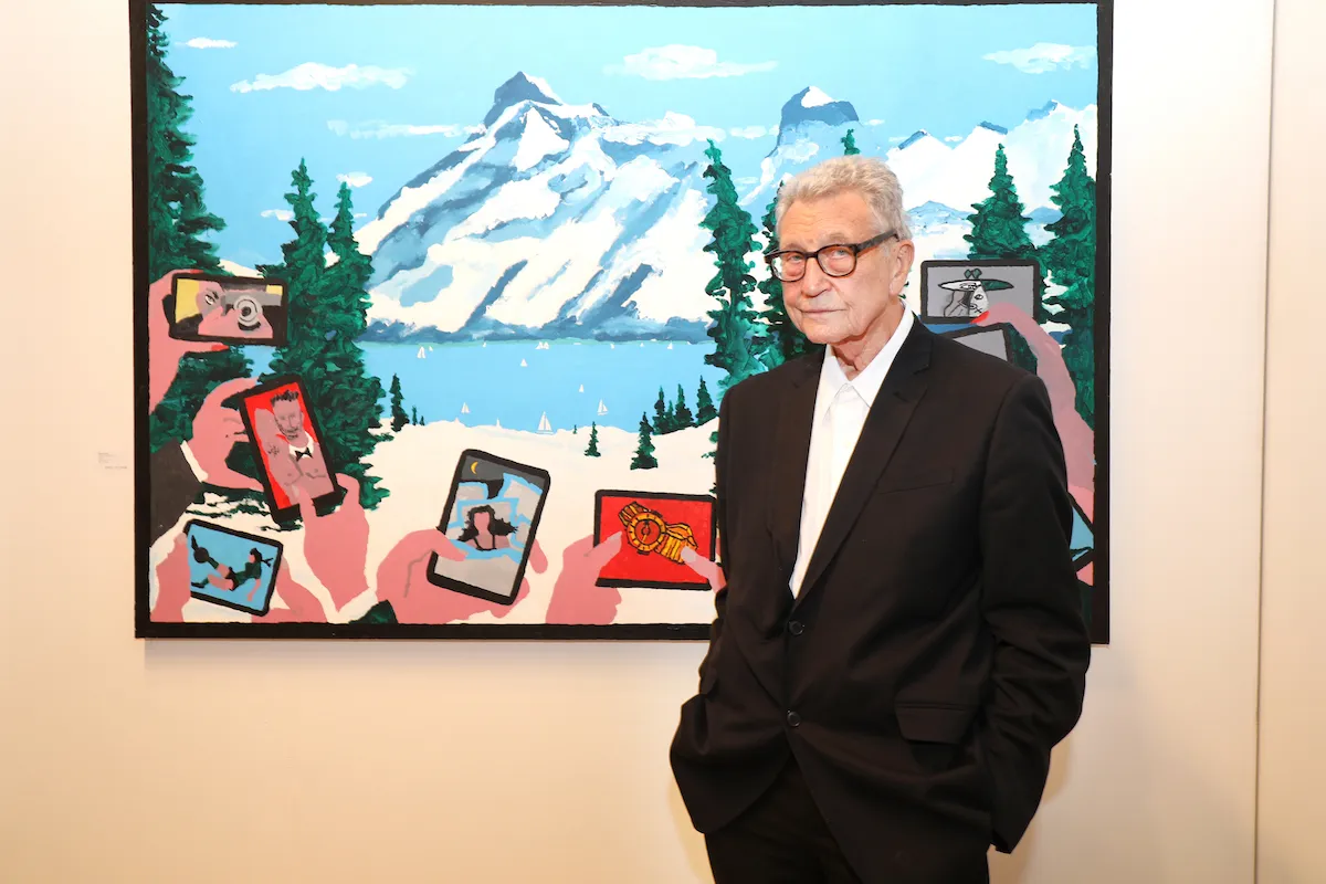 A white man in a suit beside a painting of people holding phones up to a mountain landscape. Rather than showing images of the mountain, the phones display pictures of people posing.