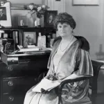 An archival black-and-white photograph of a rich white woman seated in front of a writing desk.