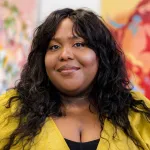 A Black woman in a mustard-colored shirt smiling before two abstract paintings.