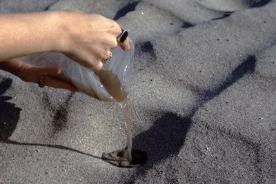 A hand pouring liquid from a plastic bag onto sand.