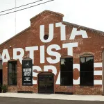 Exterior of a brick-building that had UTA Artist Space painted in white on the facade (though letters are not complete because of the architecture).