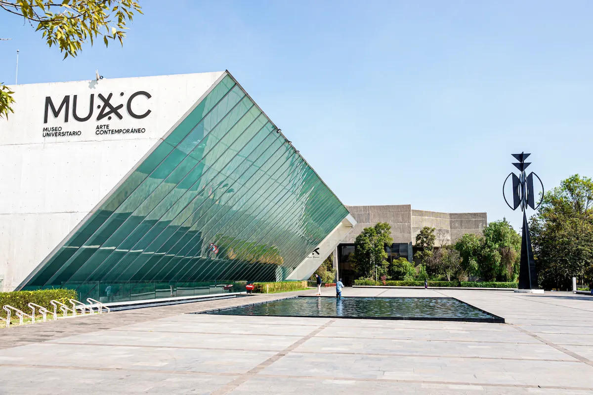 A glassy museum facade that tilts over a reflecting pool in a plaza.