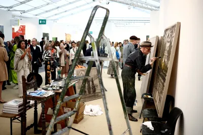 Artist Billy Childish paints on a canvas as a crowd looks on at Frieze London 2024.