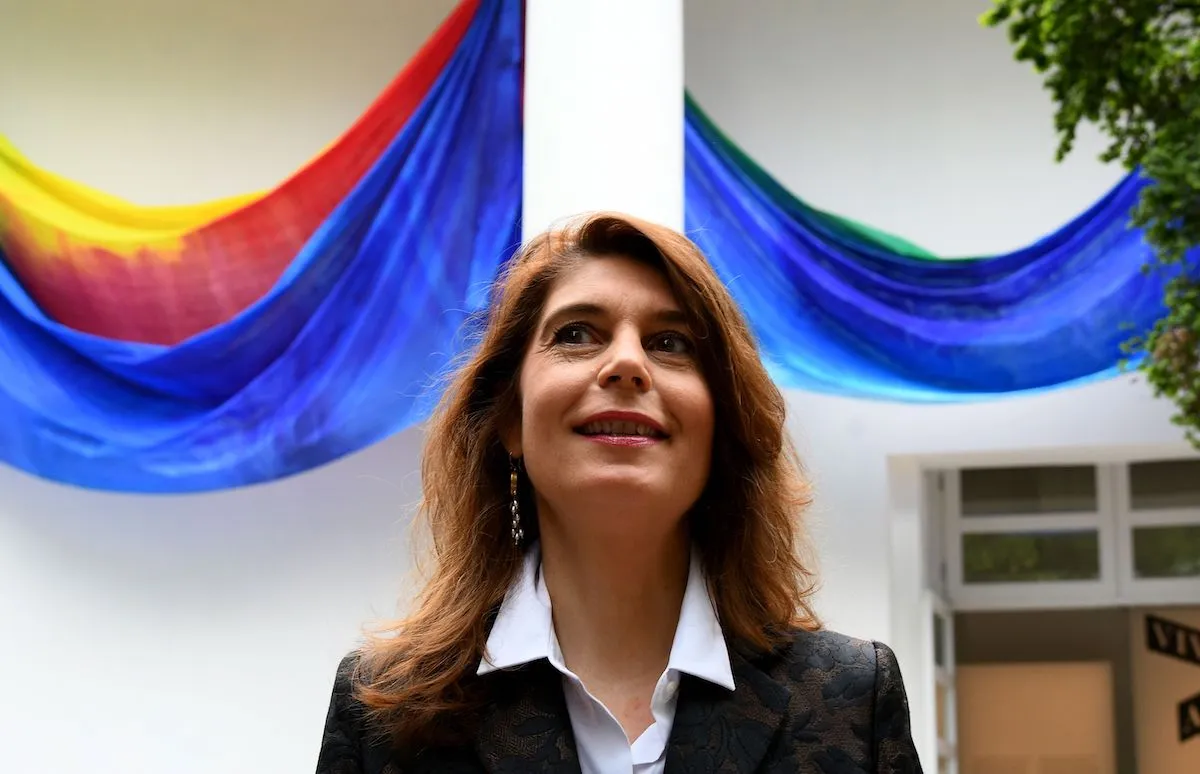 A white woman standing before an art installation composed of a hanging painted cloth.