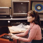 A woman in a red and white and shirt seated at a desk before two computer monitors.