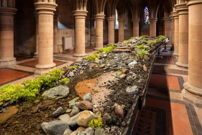 An installation consisting of rocks, earth, water, and plants on a raised platform in an architectural space with two colonnades. 