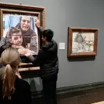 Two people pasting an image of a crying mother and child over a painting in a gallery.