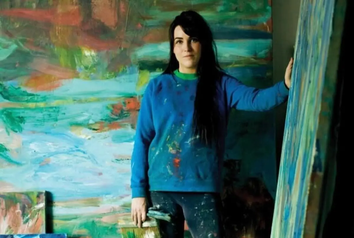 A white woman in a paint-splattered blue sweater placing one hand on an abstract canvas, with another canvas behind her.