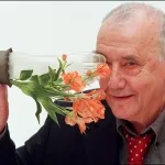 A smiling white man in a suit holding a bottle with flowers poking out of it to his face.