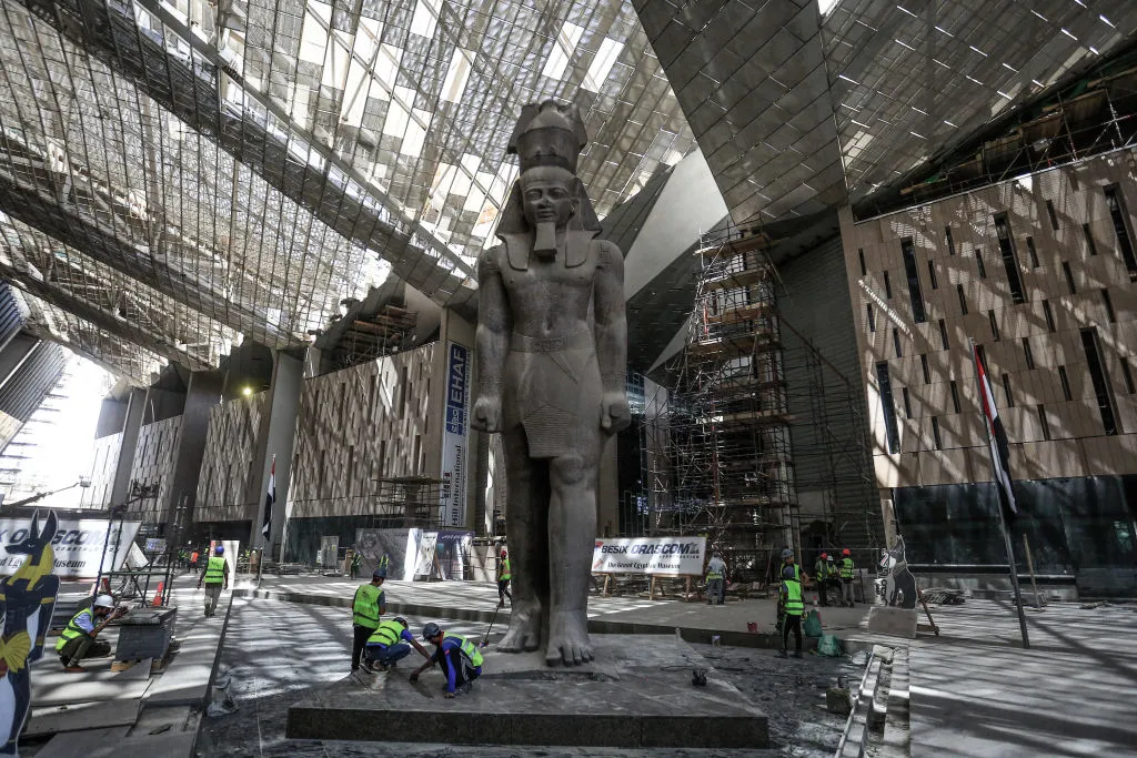 A huge statue of an Egyptian pharaoh with construction workers by its feet.