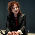 A white woman with curly red hair sitting in a chair.