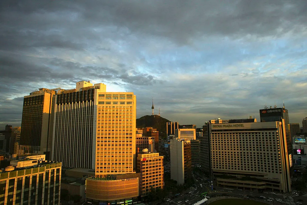 A crowded cityscape is seen from the air on a partially cloudy day that is growing dark. A tower stands on a tall hill in the distance.