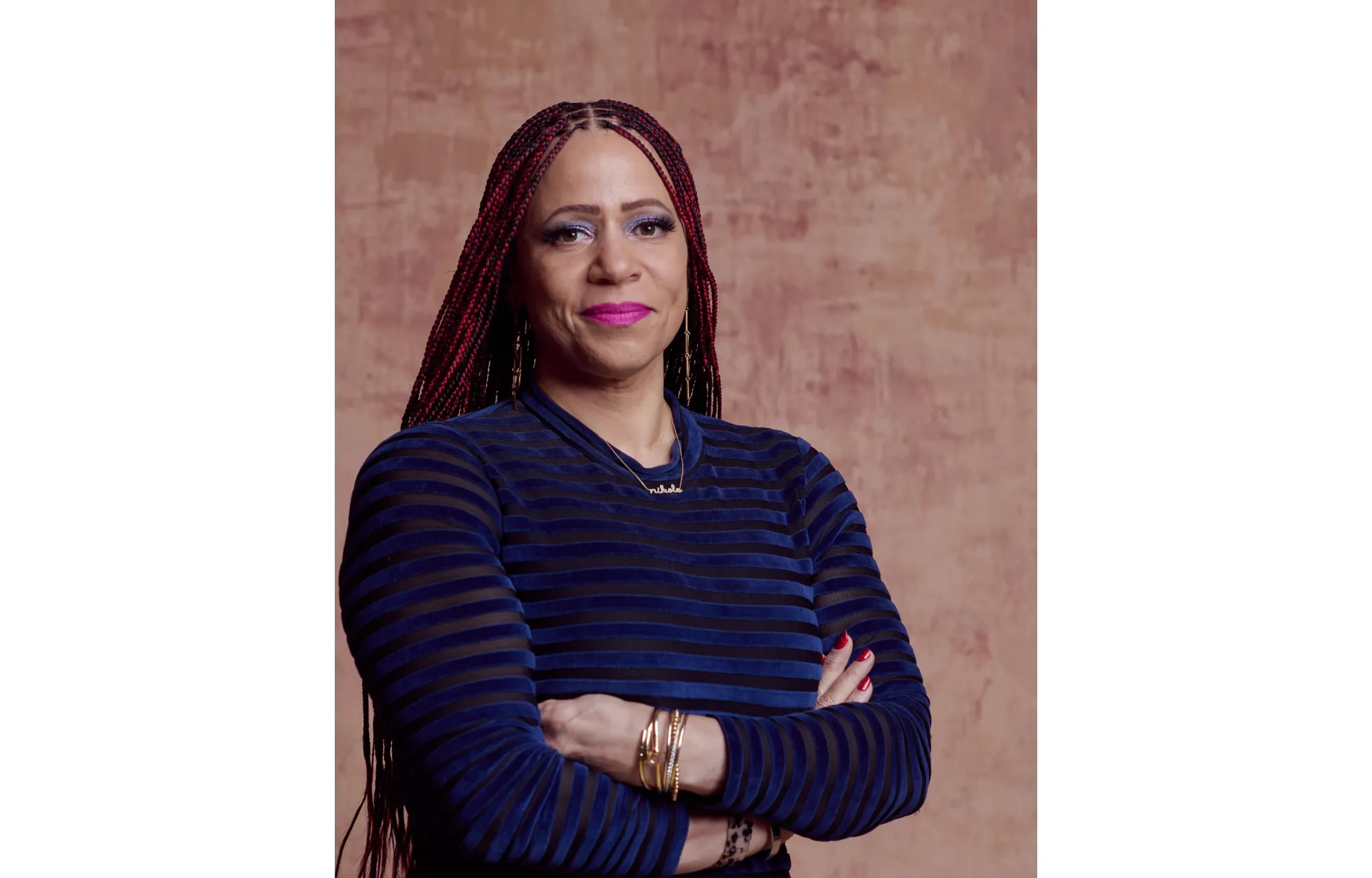 A headshot of journalist and art collector Nikole Hannah-Jones. Her hair is braided, she is wearing pink lipstick, a necklace with her name, and her arms are crossed. The Pulitzer Prize-winner recently led a tour of the 1619 Project Art Exhibition at the Brooklyn Public Library