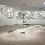 A cascading silver form in the corner of a gallery is shown amid grphite marks covering a wall. In the foreground, a linear lump of sand is punctuated with vertical rectangular mirrors.