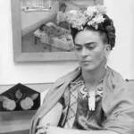 circa 1945:  Mexican painter Frida Kahlo (1907 - 1954) sits with her arms folded, looking down, in front of one of her paintings and a wooden bird cage. She wears flowers in her hair and a wooden necklace.  (Photo by Hulton Archive/Getty Images)