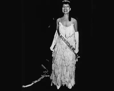 A Black woman wearing a dress made out of gloves with a sash reading 