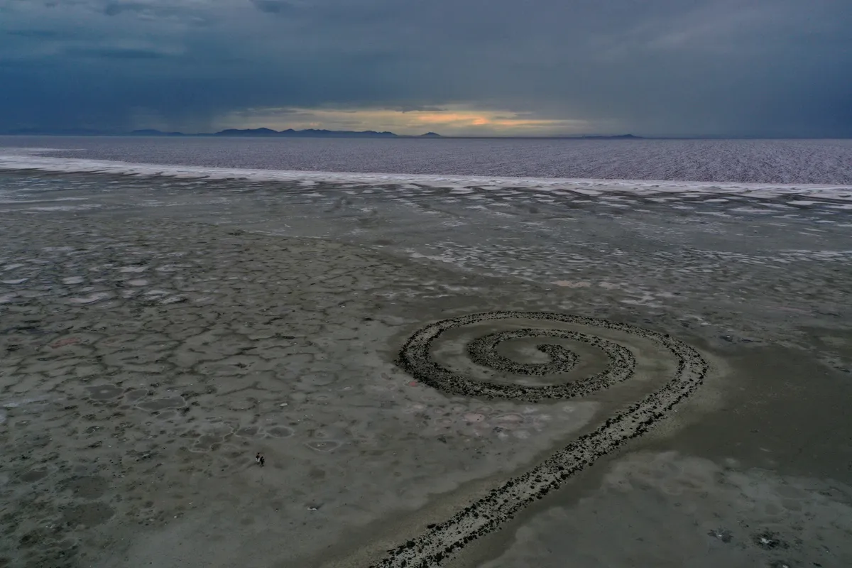 A spiral form appears in the muddy foreground with a white-edged body of water beyond it and a faint sunset peeks through a dark clouded sky.