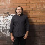 A man pictured against a graffitied brick wall.